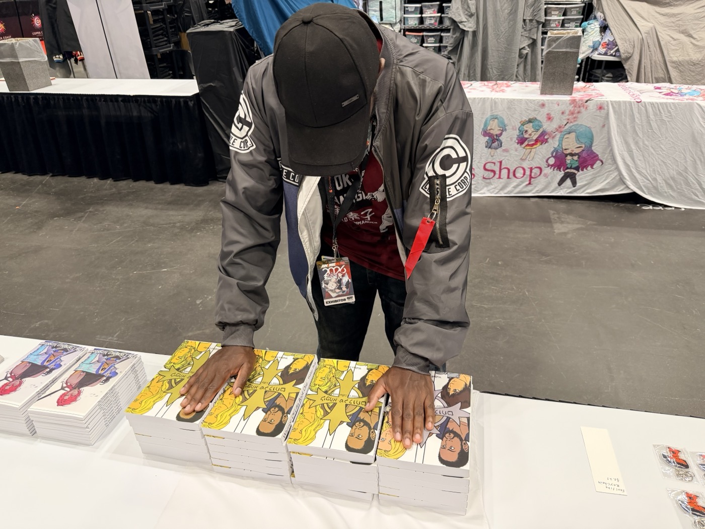 A team member leaning over the booth table, squaring up stacks of Warrior Of Lifta before the show opens.
