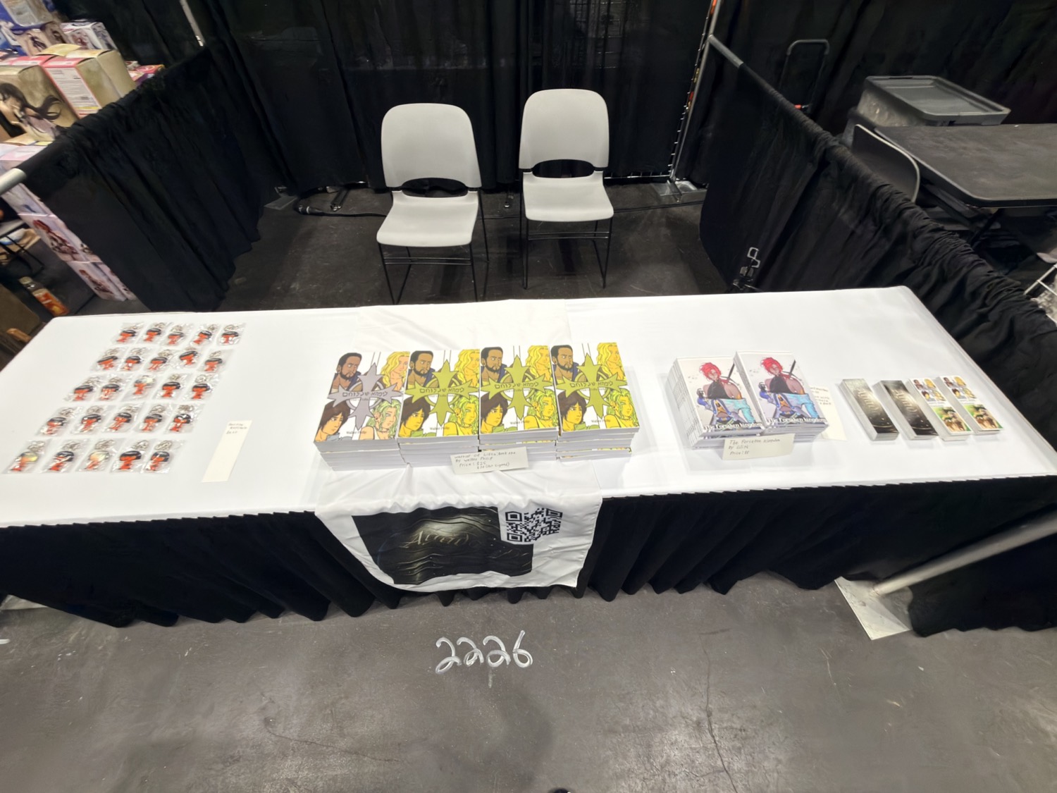 The Agada Publishings booth table laid out with book stacks, art prints, and enamel pins on a white cloth.